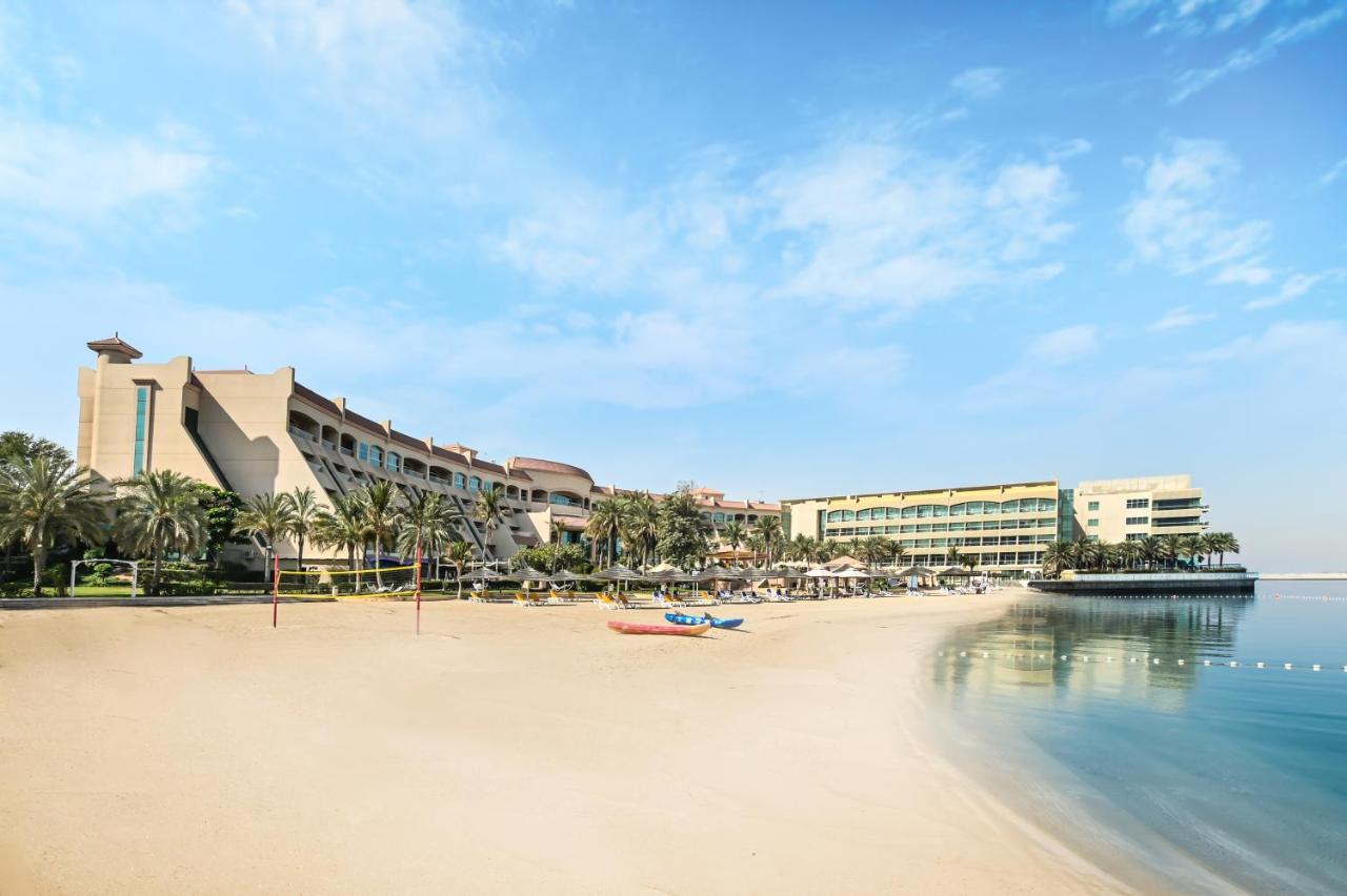 فندق شاطئ الراحة أبو ظبي المظهر الخارجي الصورة The photo depicts a serene beach scene with a clear blue sky. In the foreground, there is a sandy beach that gently slopes down to calm water. On the left side, several palm trees provide a tropical atmosphere. In the background, there are two large 
