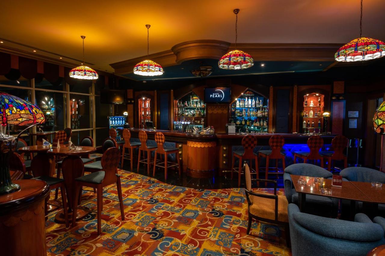 فندق شاطئ الراحة أبو ظبي المظهر الخارجي الصورة The photo shows a cozy bar interior, featuring a well-stocked bar area with various bottles displayed on shelves. The bar is illuminated by colorful stained glass light fixtures, creating a warm and inviting atmosphere. The floor is covered with a vi