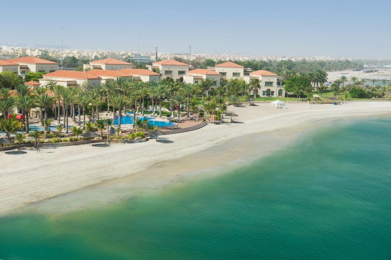 فندق شاطئ الراحة أبو ظبي المظهر الخارجي الصورة The photo depicts a serene beach scene with a clear turquoise sea and a sandy shoreline. In the background, there are several buildings featuring a Mediterranean architectural style with red-tiled roofs. The area is adorned with palm trees and outdoo