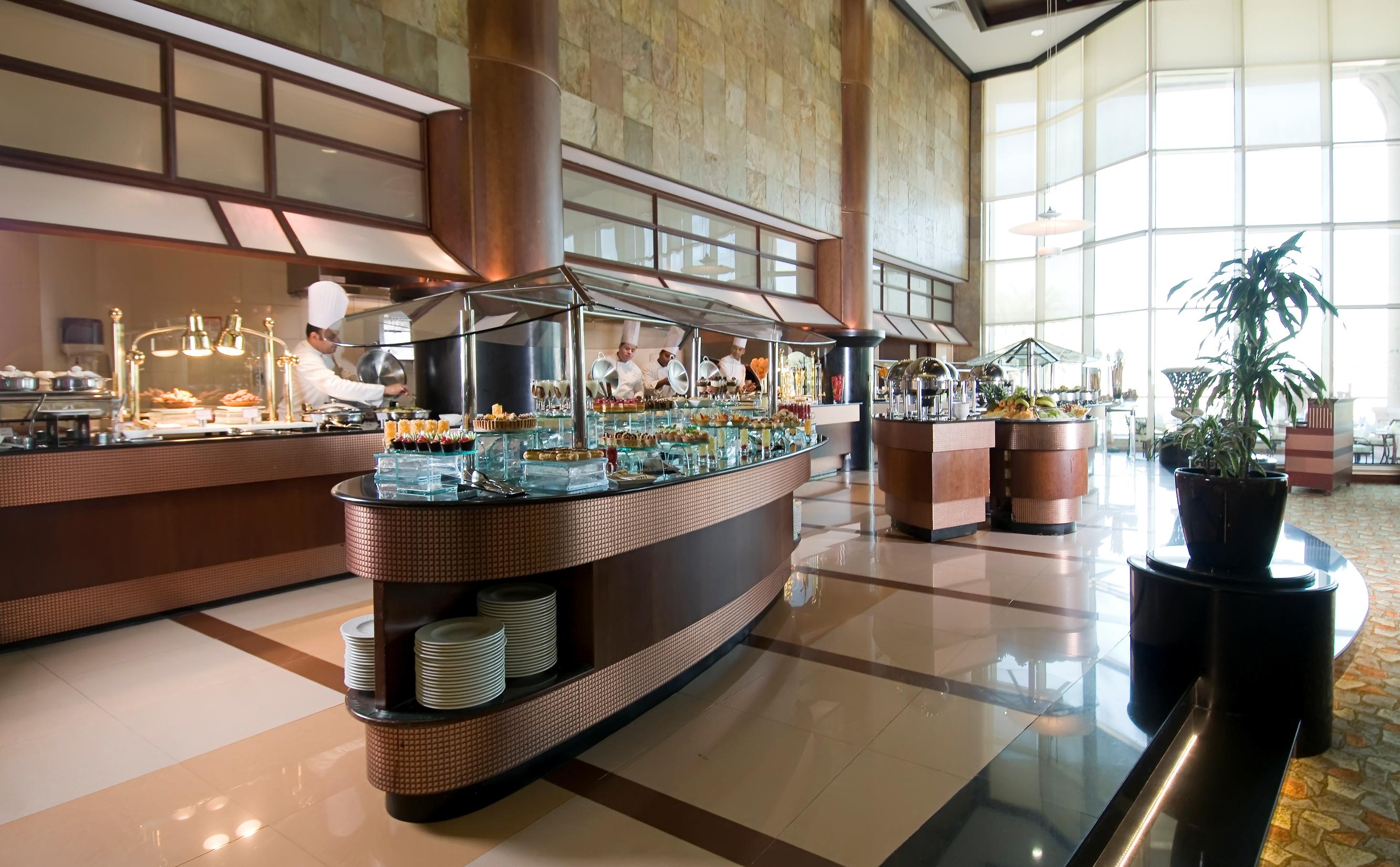 فندق شاطئ الراحة أبو ظبي المظهر الخارجي الصورة The photo shows a spacious buffet area in a restaurant or hotel. There are multiple food stations with glass displays showcasing a variety of dishes. Chefs can be seen in the background, preparing food. The decor features large windows allowing natur