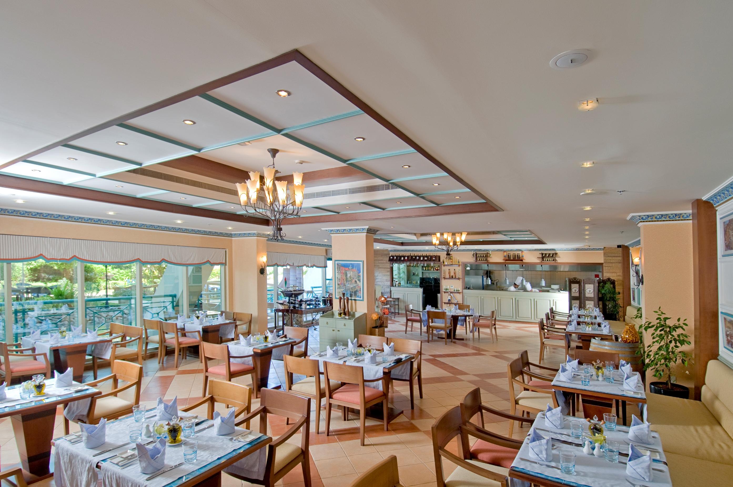 فندق شاطئ الراحة أبو ظبي المطعم الصورة The photo shows the interior of a restaurant. It features a spacious dining area with multiple tables neatly arranged, each set with tableware and cloth napkins. The decor includes a decorative ceiling with lighting fixtures, and large windows that p