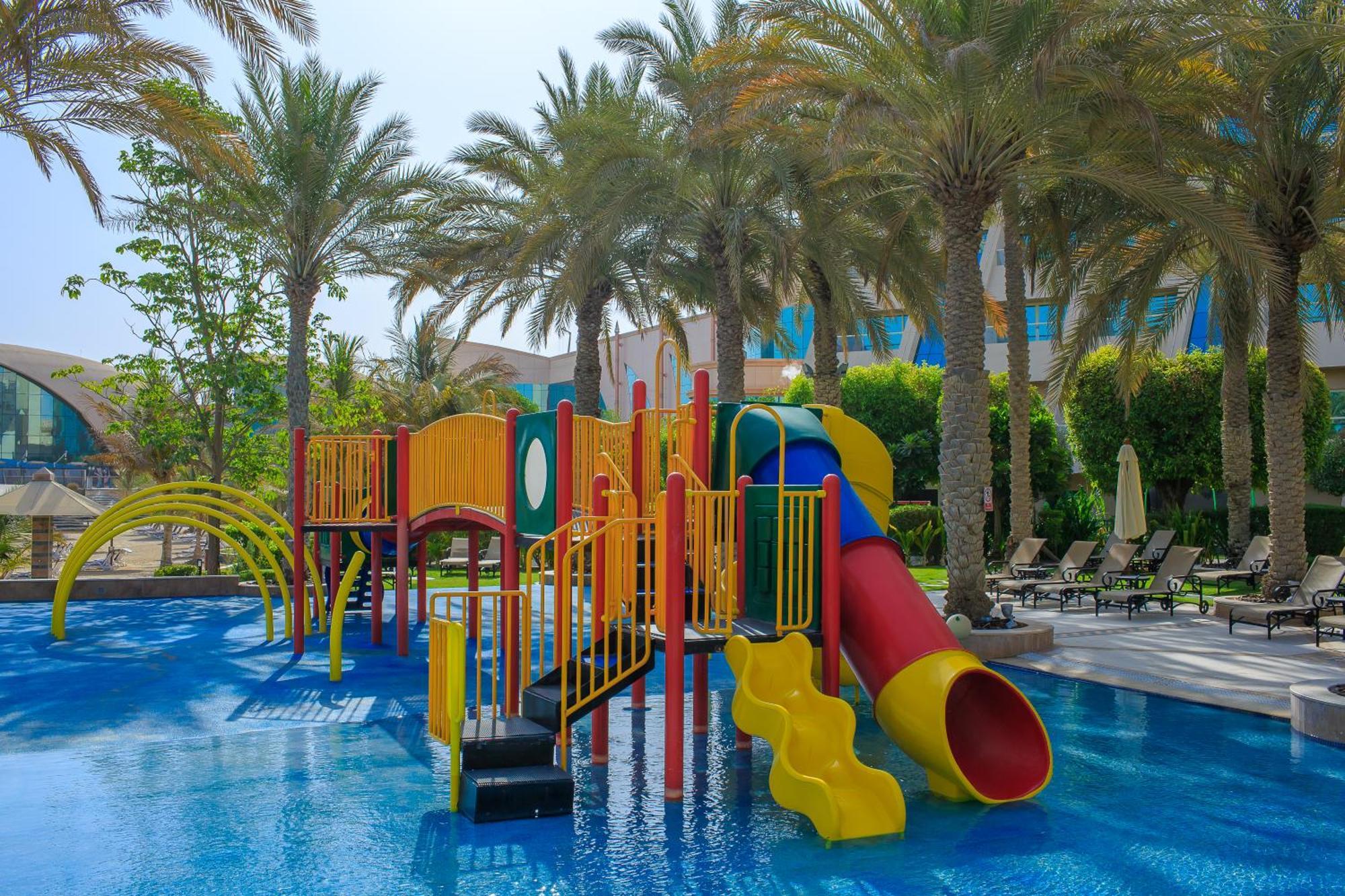 فندق شاطئ الراحة أبو ظبي المظهر الخارجي الصورة The photo shows a colorful water play area located in a pool setting. It features a variety of play structures, including slides in multiple colors, such as yellow, red, and blue. Surrounding the play structure are palm trees, creating a tropical atm