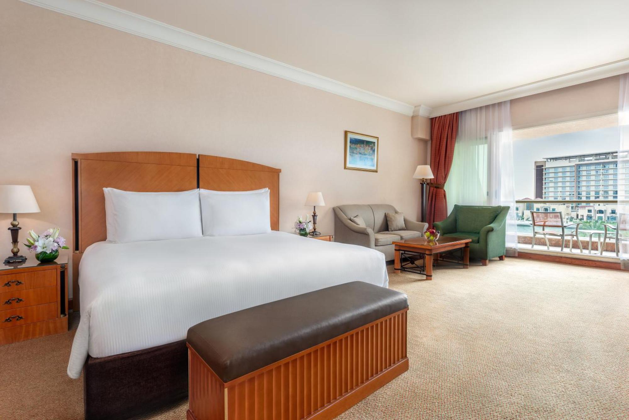 فندق شاطئ الراحة أبو ظبي المظهر الخارجي الصورة The photo shows a well-furnished hotel room featuring a large bed with white linens and a wooden headboard. There's a brown bench at the foot of the bed. To one side, a sitting area includes a sofa and a coffee table, complemented by two lamps. Large
