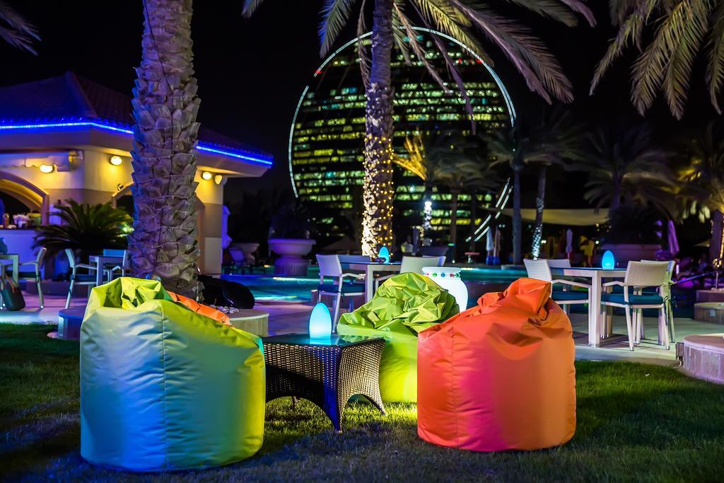 فندق شاطئ الراحة أبو ظبي المظهر الخارجي الصورة The photo shows a vibrant outdoor lounge area at night. In the foreground, there are colorful bean bag chairs—one in bright green, one in yellow, and one in orange. A small table sits between the bean bags. In the background, illuminated palm trees a