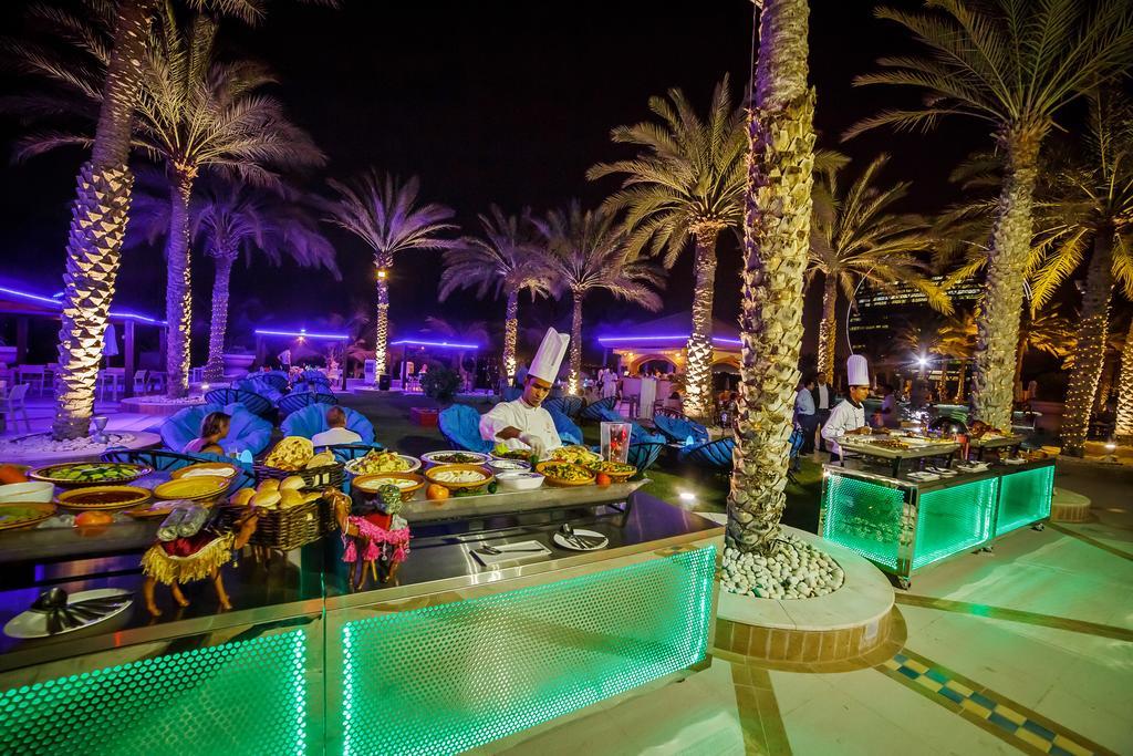 فندق شاطئ الراحة أبو ظبي المظهر الخارجي الصورة The photo depicts an outdoor dining area at night, featuring several chefs in white uniforms and hats working at a food station. The setting is illuminated by colorful lights, with glowing green accents on the serving tables. Surrounding the area are